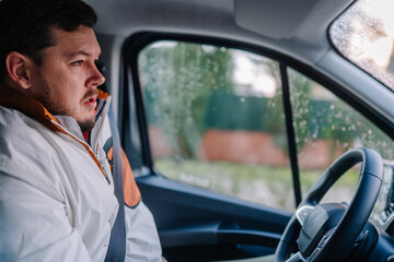 Male driver wearing a white jacket sits inside a vehicle, looking out the window with raindrops on the glass, showcasing an urban environment in the background