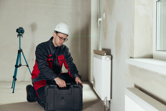 Technician Kneeling Next To Tool Case With Laser Level And Radiator In Background. Construction Workflow In Raw Interior.