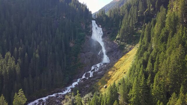 Cinematic aerial drone footage of Krimml Waterfalls in the Austrian Alps on a sunny summer day. Powerful big streams of water plunge down rugged cliffs, framed by lush green pine forest and mountains
