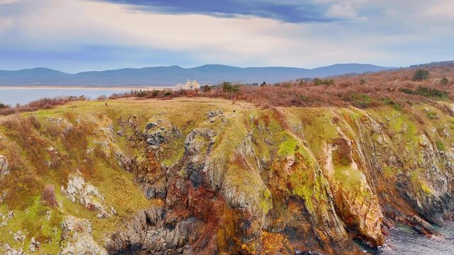 High rocky cliff overlooking clear turquoise sea water. Vertical view of layered rock formations with moss and grass above clear blue water.