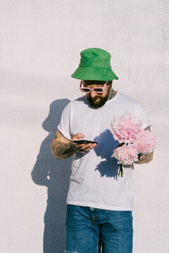 Man in green hat holds phone and peonies against white wall