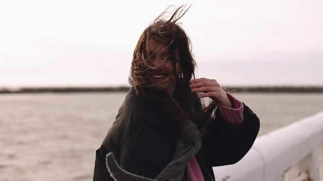 Young woman looking around then flirting with camera, laughing and smiling in warm sunset light by the windy seaside, hair blowing in strong wind, city and ocean view, black puffer jacket pink sweater