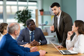 Man stands in front of business colleagues, tells report, proves vision of situation, defends way to solve problems. People are sitting at table, and participant is standing up and took floor. . © JackF