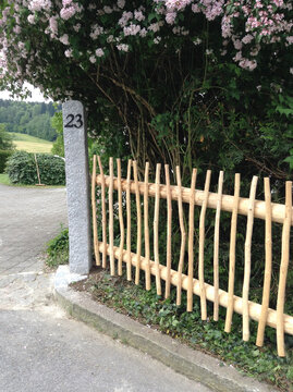 &ldquo;A peaceful country scene with a rustic wooden fence and a stone gatepost marked with the house number 23, surrounded by blooming flowers and green hills