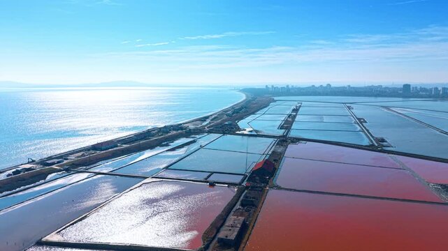 Wide aerial view of salterns and sea under clear sky. High angle perspective of salt evaporation fields stretching towards the sea and coastal city.