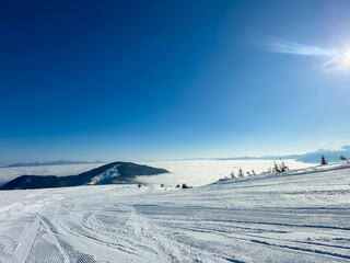 Snow-covered mountain slopes under clear blue sky with clouds below and bright sunlight shining...