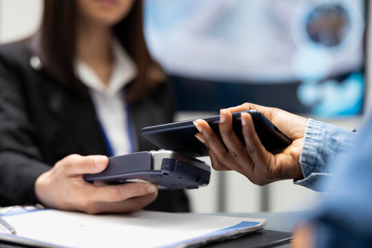 Patient using smartphone for cashless medical payment as clinic receptionist processes transaction with card reader. Black individual pays hospital fees, tapping mobile device on pos machine.