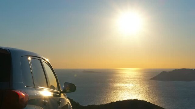 Car parked on a cliff overlooking the sea at sunset. Golden sunlight reflecting on a silver car parked on a scenic coastal road overlooking a beautiful seascape with islands on the horizon