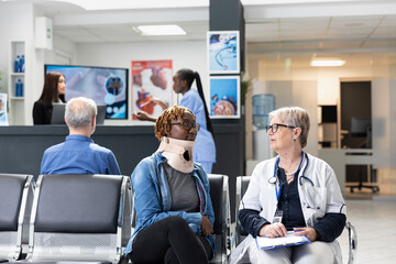 Fototapeta premium Attentive doctor providing friendly assistance to patient with neck brace seated in hospital waiting room. Retired female physician wearing lab coat reassuring injured black woman of speedy recovery.