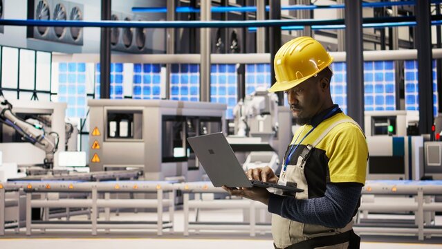Technician in solar panels factory oversees robot assembly lines using laptop, ensuring quality control. Industrial plant worker monitoring automated conveyor systems using notebook, camera A