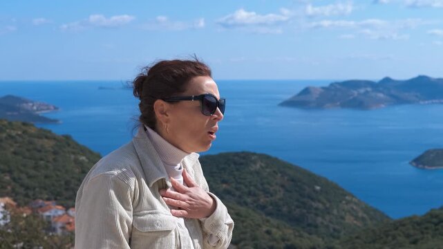 Woman with shortness of breath using asthma inhaler outdoors. Mature woman experiencing breathing difficulties and an asthma attack, using a preventer inhaler while standing on a mountain with a sea