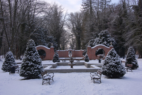 Park dworski w mieście Iłowa w zimowy dzień. Faliste, ażurowe, ceglane ściany, noszące nazwy bram księżycowych wraz ze znajdującymi się w sąsiedztwie nieckami fontann. Ziemię pokrywa śnieg.