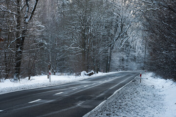 Asfaltowa droga w zimowy dzień. Jezdnia jest czarna, po obu stronach jest wysoki las. Ziemia i gałęzie drzew pokrywa warstwa śniegu. © boguslavus