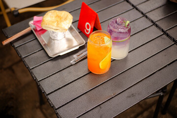 A refreshing orange and purple mocktail is served with golden cookies on a black folding table number 9 in an outdoor cafe © Khoirul