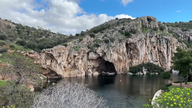Iconic Vouliagmeni lake panoramic view with towering limestone cliffs, dramatic cave arch reflecting in calm turquoise water and lush mediterranean shrubs in foreground, Greece
