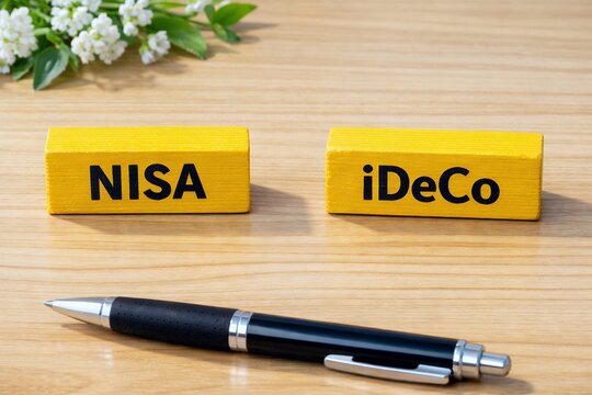 NISAやiDeCoのロゴが印字された黄色い積み木と黒いボールペンがある資産運用のイメージ