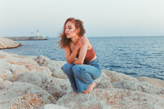 Woman Sitting on Rocky Shore Near Lighthouse During Sunset