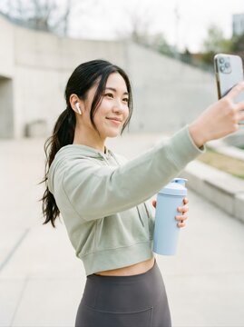 young sporty woman taking selfie on smartphone after workout holding shaker bottle outdoors. smiling and listening to music with earbuds. ugc fitness content and healthy lifestyle concept