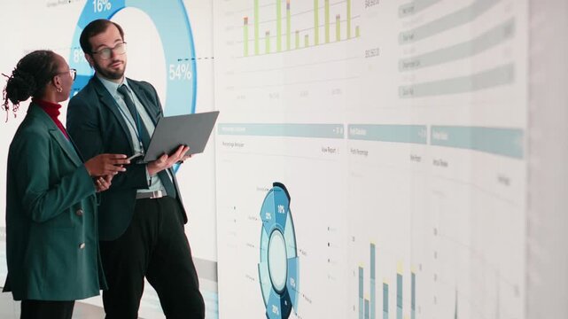 Corporate managers evaluate fiscal performance metrics on digital wall, collaborating. Diverse executive team discusses quarterly earnings report and shareholder equity charts during boardroom meeting