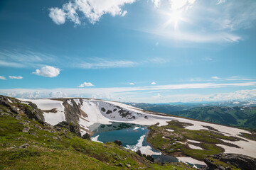 Scenic sunlit landscape with alpine lake in rocky snowy cirque near stone hill top in sunny day during thaw. Ice floats in mountain lake among rocks with view to forest mountain range under bright sun © Daniil