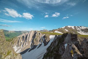 Aerial dizzying view to rocky cliff between sharp rocks under clouds in blue sky. Vertigo scenery with sheer crags above precipice, snowy ridge and mountain top far away in cloudy changeable weather. © Daniil
