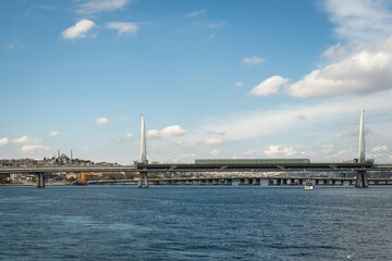 Fototapeta premium Golden Horn Metro Bridge (Halic Metro Koprusu) spanning the Golden Horn with calm water, skyline, and a small boat under a blue sky in Istanbul, Turkey