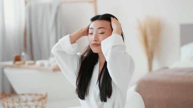 Indoors portrait of young well groomed asian woman preening at bathroom, fixing her hairstyle and smiling, looking at herself, slow motion