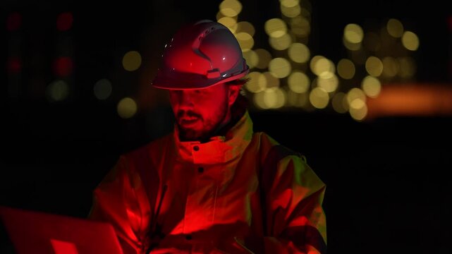 Focused male industrial engineer working late night shift under red emergency warning light. Professional worker wearing safety helmet inspecting factory operation outdoor.