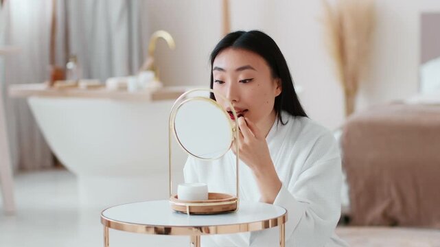 Young beautiful asian woman applying red lipstick on lips, making bright make-up, sitting at blurred bathroom interior, slow motion, free space
