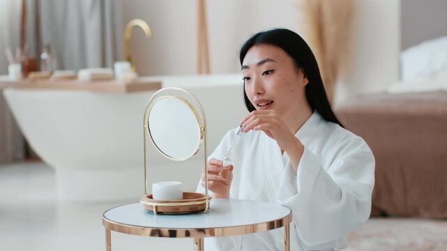 Skin care and vitamization. Young pretty asian lady applying nourishing serum on face looking at mirror, making facial pampering rituals ta bathroom at home, slow motion, free space