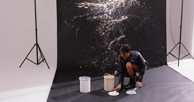 Asian woman 20s jacket opening buckets flicking paint making splatter wall in studio, copy space