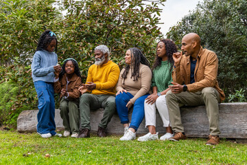 African American family with kids and seniors sitting on log in park sharing smartphone in sweaters