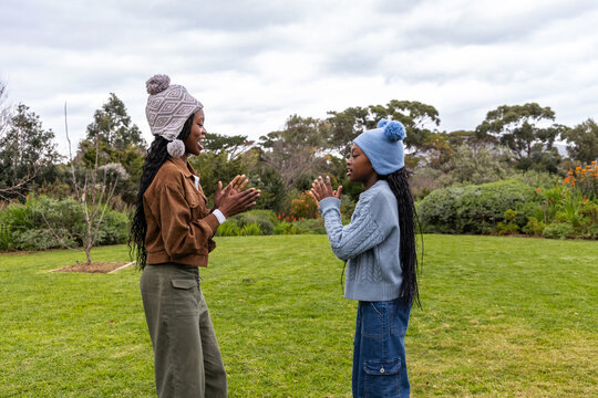 Two African American preteen girls playing clapping game on lawn, wearing pom-pom hats and sweaters