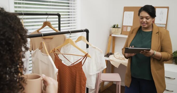 Diverse female colleagues browsing rack, swiping tablet for garment selection in office, copy space