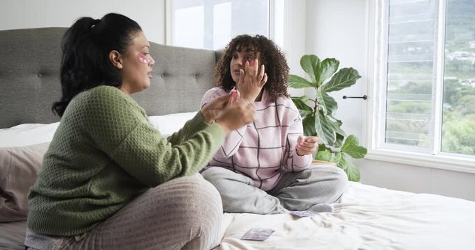 Mother and daughter on bed scooping eye patches from bowl, pressing patches under eyes for skincare