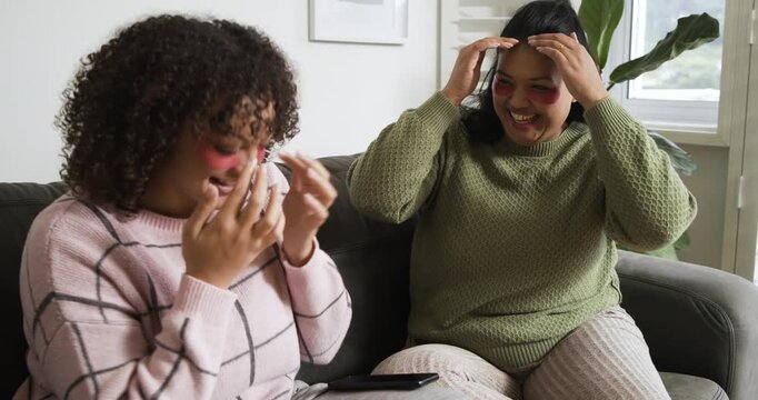 Diverse female friends applying hydrogel patches on sofa tapping phone and playfully taking selfies