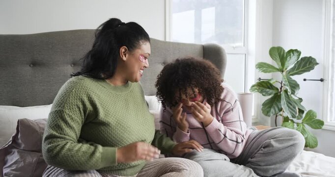 Grabbing patches for skincare, African American mother and daughter applying them, laughing on bed
