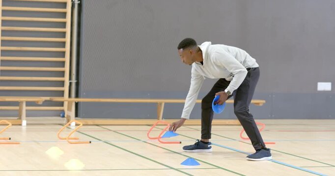 African American male coach entering gym placing discs by hurdles for volleyball drill