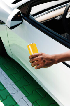 Hand holding yellow drink can beside white parked car