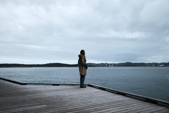 Person Standing on Wooden Deck Overlooking Calm Ocean on a Cloudy Day