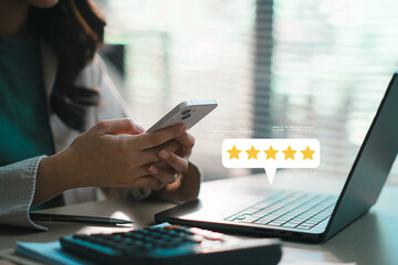 Woman using smartphone with five star rating feedback icon on screen, writing online review while working on laptop, customer satisfaction survey, digital marketing and service evaluation