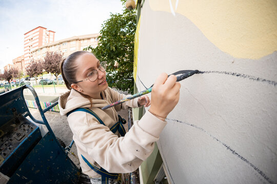 Female artist painting a mural on a building wall using a lift