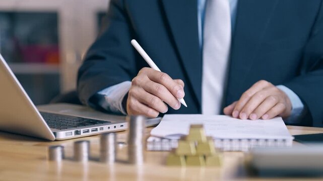 Businessman counting coins and gold bars, reviewing financial data, and signing business report for investment approval and final agreement.