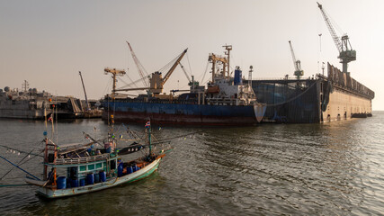 Traditional wooden Thai fishing boat with massive industrial and cargo terminal, Contrast and coexistence between local livelihoods and growth modern heavy industry.