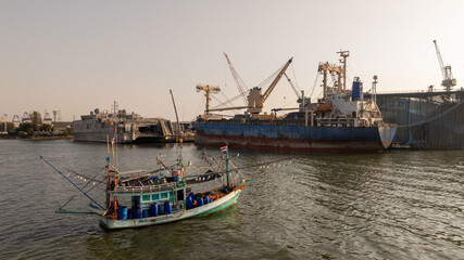 Traditional wooden Thai fishing boat with massive industrial and cargo terminal, Contrast and coexistence between local livelihoods and growth modern heavy industry.