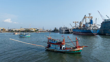 Traditional wooden Thai fishing boat with massive industrial and cargo terminal, Contrast and coexistence between local livelihoods and growth modern heavy industry.