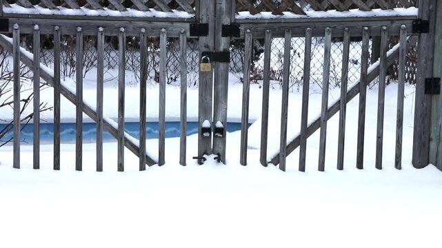 Snow-covered wooden gate with a padlock, leading to a frozen pool, surrounded by a tranquil winter landscape under soft, diffused light.