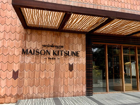  A modern storefront featuring the Maison Kitsune brand. The building has a distinctive patterned facade with warm tones and a large glass entrance