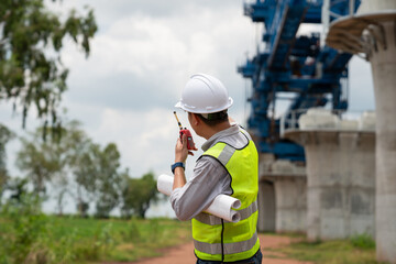 The survey team is operating a theodolite while referencing road construction plans. Civil engineers are taking measurements with surveying equipment at the construction site.