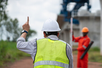The survey team is operating a theodolite while referencing road construction plans. Civil engineers are taking measurements with surveying equipment at the construction site.
